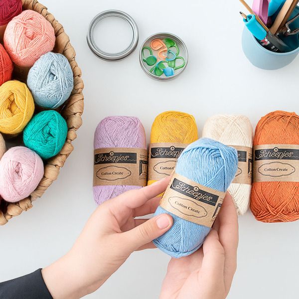 scheepjes cotton create katoengarens in diverse kleuren, creatief uitgestald op een werktafel met accessoires en handen die een bol vasthouden